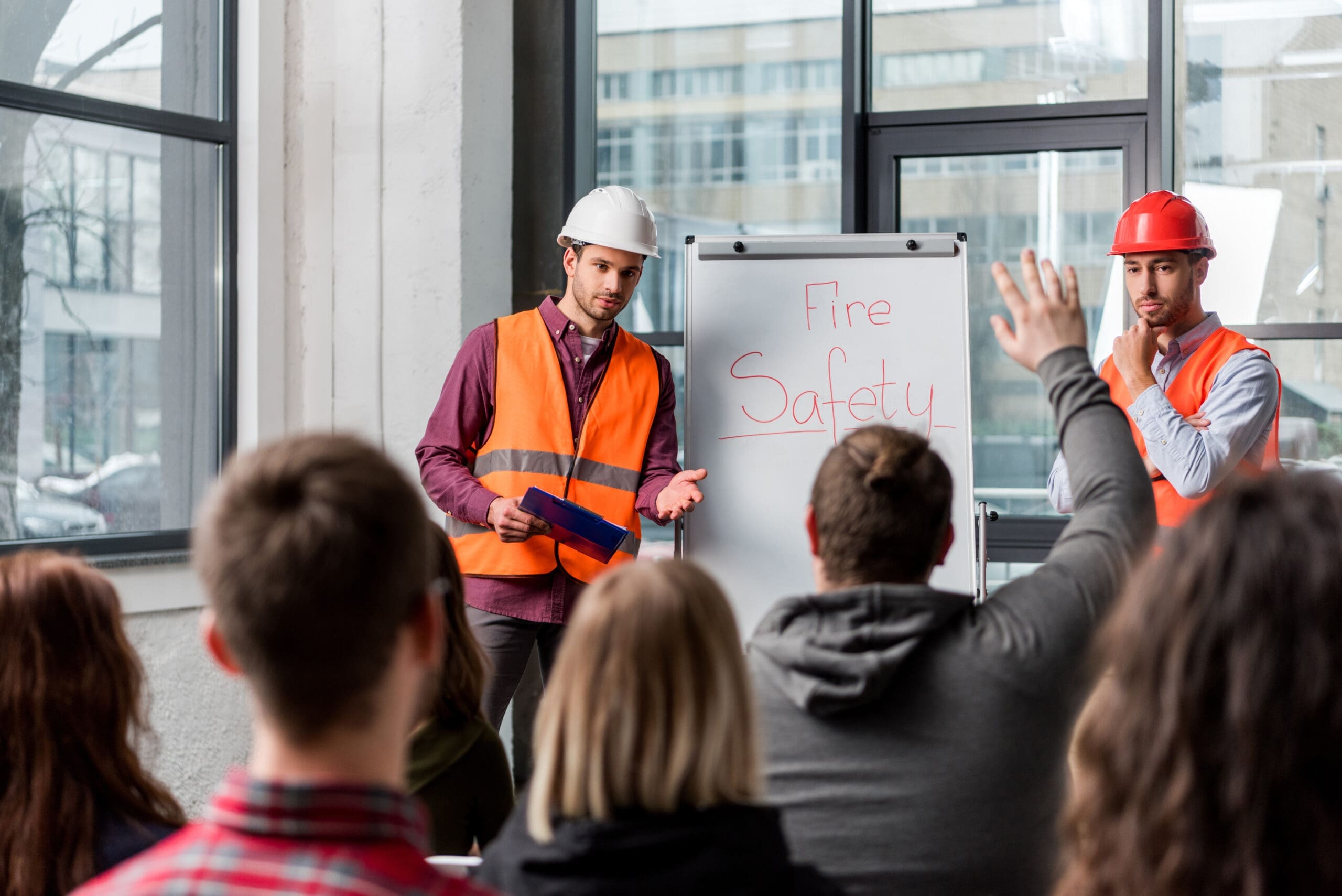 Selective focus of handsome firemen in helmets giving talk on briefing near white board with fire safety lettering and looking at man raising hand