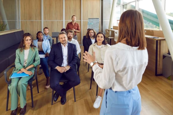 Confident female business coach teaches small group of people new business knowledge. People are sitting on chairs in office of business center listening to woman standing with her back to camera.