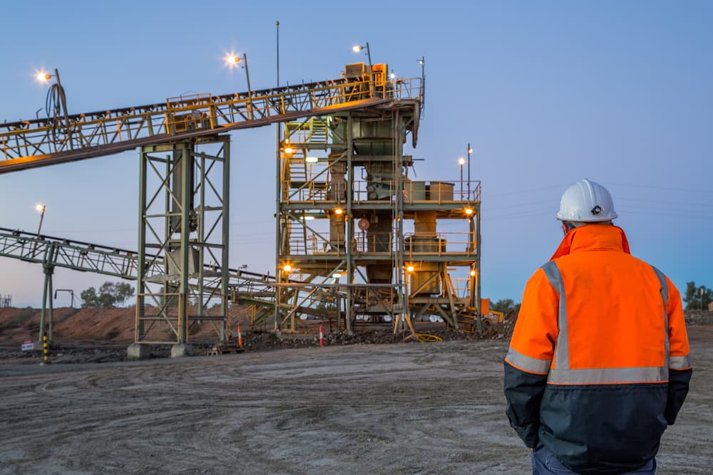 A miner in high vis workwear stands looking at a big processing plant in the dusk