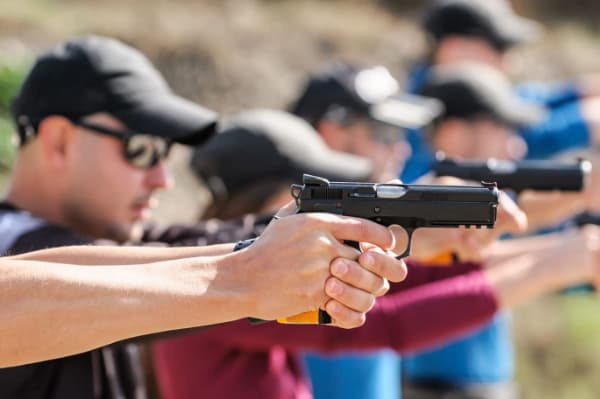 A row of people in the sunshine all pointing handguns off the the right.